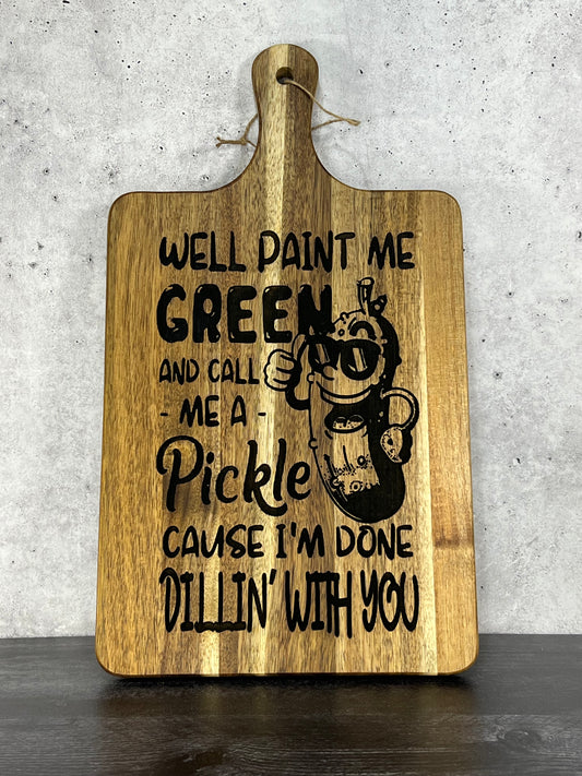 Acacia wood cutting board measuring 16" x 9", laser engraved with a cartoon pickle wearing sunglasses and the phrase “Well paint me green and call me a pickle ‘cause I’m done dillin’ with you,” displayed upright on a dark table against a concrete wall background.
