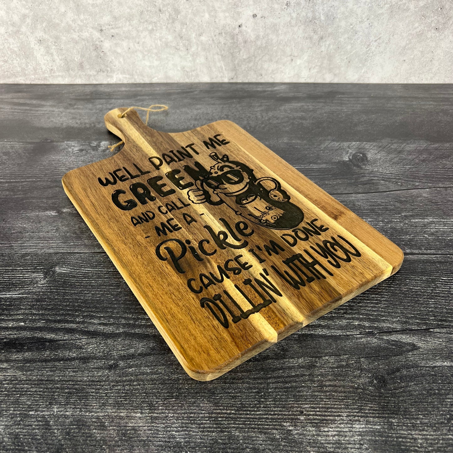Acacia wood cutting board measuring 16" x 9", laser engraved with a cartoon pickle wearing sunglasses and the phrase “Well paint me green and call me a pickle ‘cause I’m done dillin’ with you,” displayed upright on a dark table against a concrete wall background.