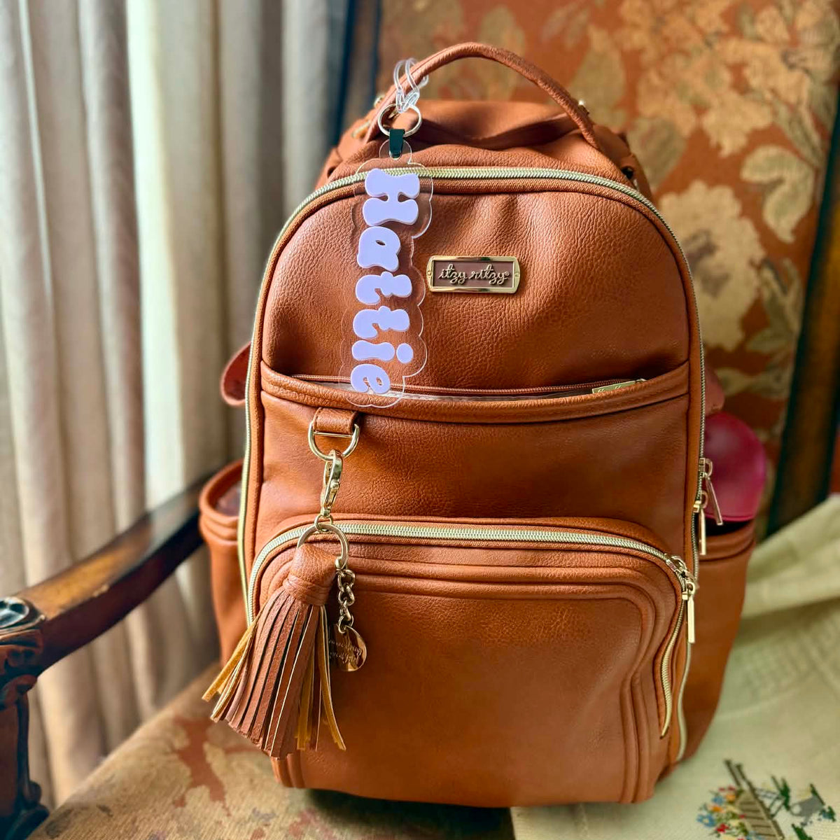 A brown faux-leather backpack with multiple zippered compartments and a tassel charm, resting on an ornate patterned armchair next to a curtain.