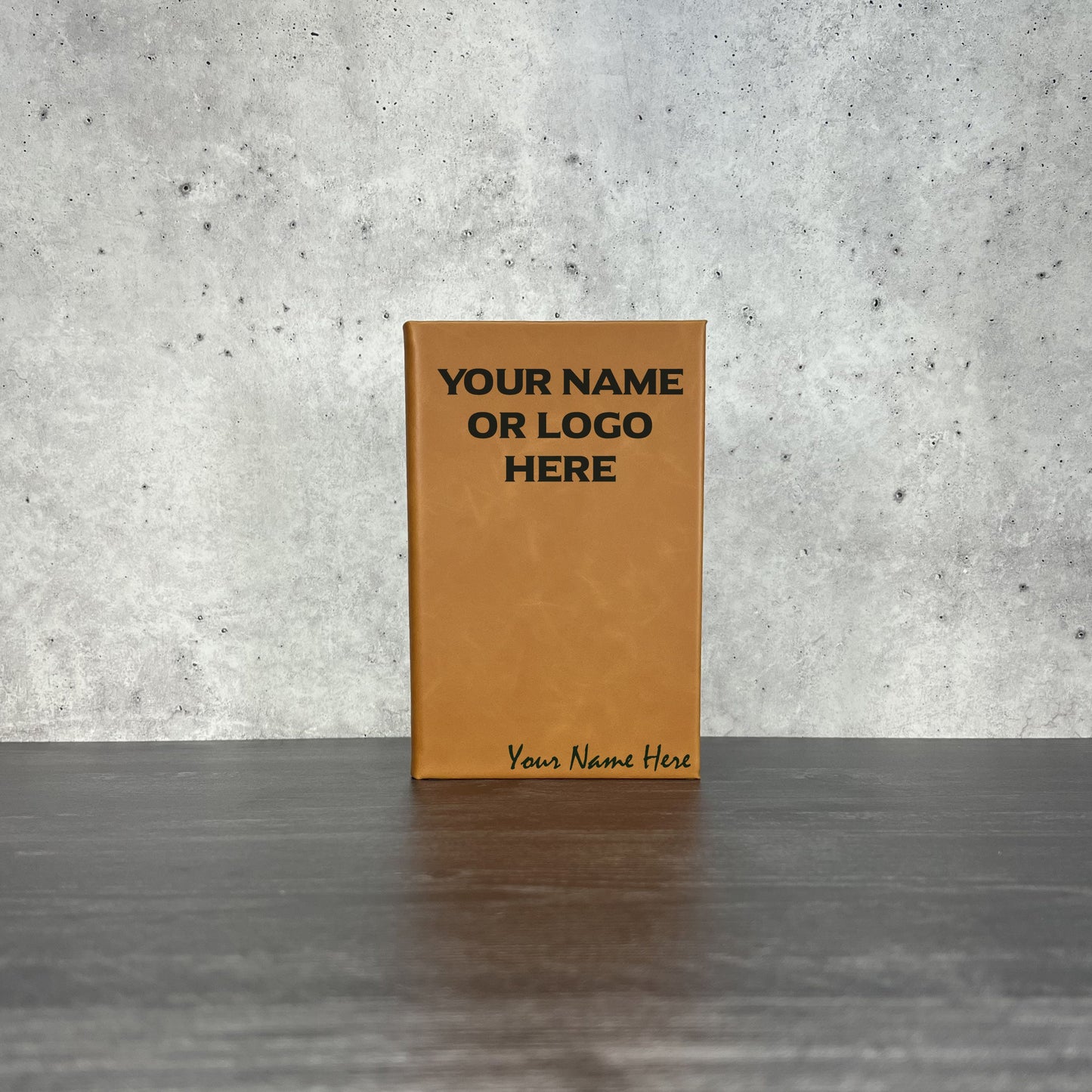 Brown leather notebook with customizable text on a gray surface and concrete wall background