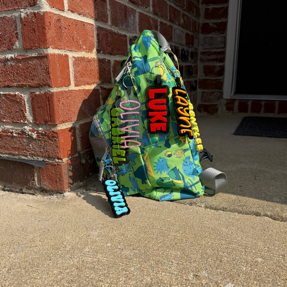 A green and blue camouflage sling backpack with colorful graphic text patches, resting on a concrete step next to a red brick wall.
