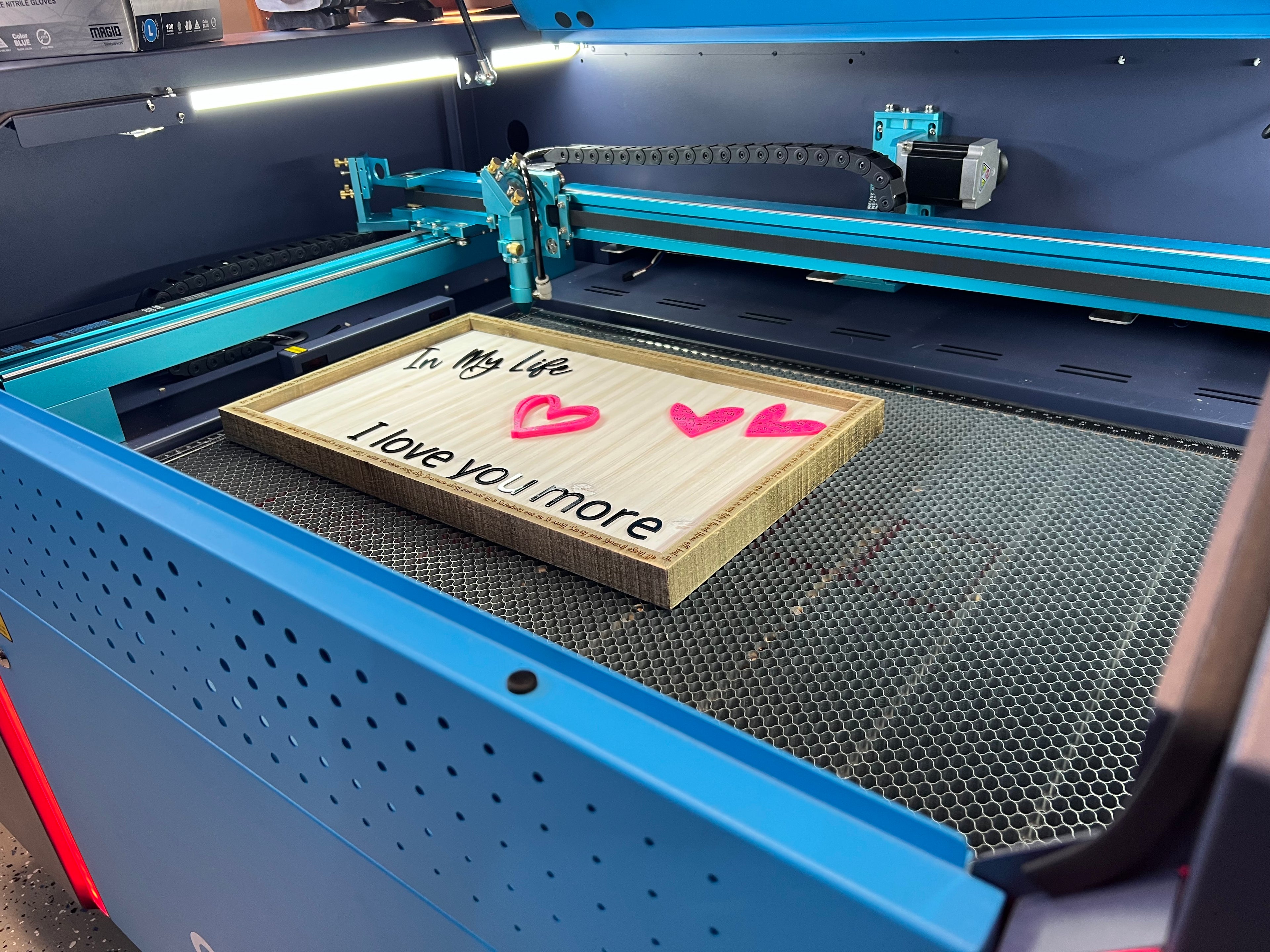 Laser cutting machine with a wooden plaque featuring 'In my life I love you more' text and hearts.