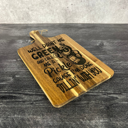 Acacia wood cutting board measuring 16" x 9", laser engraved with a cartoon pickle wearing sunglasses and the phrase “Well paint me green and call me a pickle ‘cause I’m done dillin’ with you,” displayed upright on a dark table against a concrete wall background.