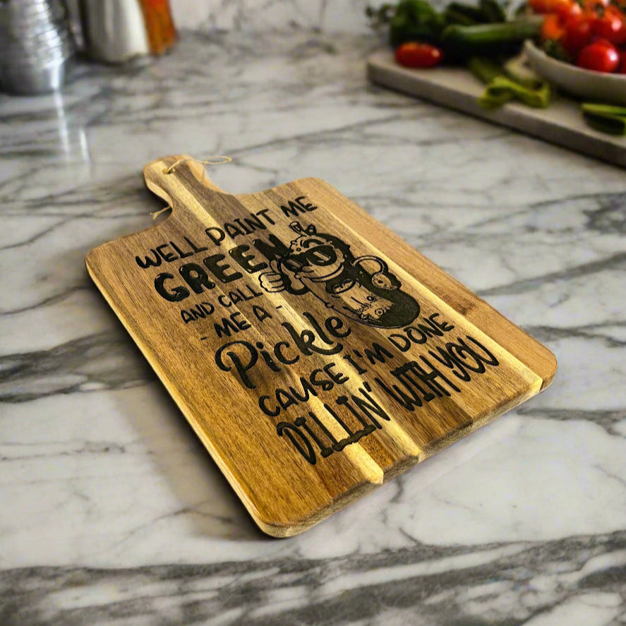 Acacia wood cutting board measuring 16" x 9", laser engraved with a cartoon pickle wearing sunglasses and the phrase “Well paint me green and call me a pickle ‘cause I’m done dillin’ with you,” displayed upright on a dark table against a concrete wall background.