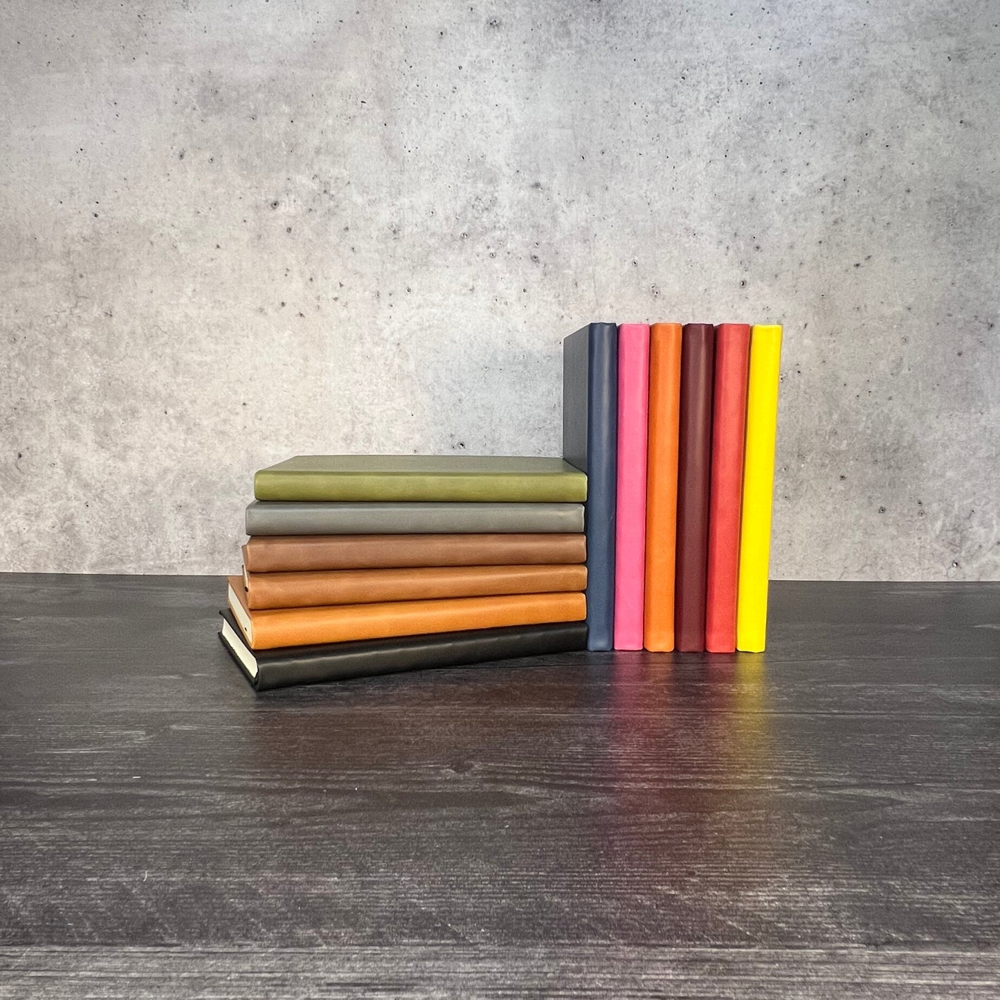 Colorful leatherette journals stacked on a wooden surface with a concrete wall background