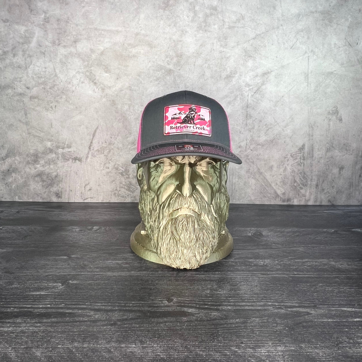 A dark gray and pink trucker hat with a graphic patch, displayed on a decorative green skull bust against a textured gray wall and dark wood table.