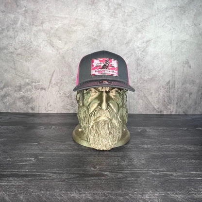 A dark gray and pink trucker hat with a graphic patch, displayed on a decorative green skull bust against a textured gray wall and dark wood table.