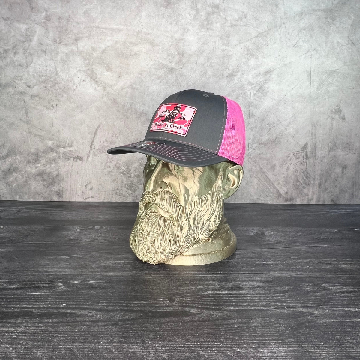 A black and pink trucker hat with a red and white front patch logo, displayed on a green mannequin head against a gray textured wall and dark wood floor.