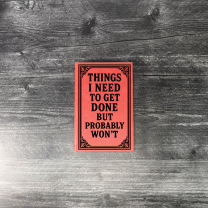 Red journal with text 'Things I need to get done but probably won't' on a wooden surface.